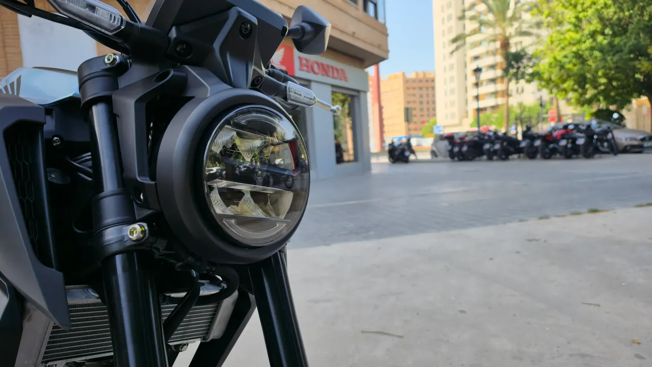 Faro delantero LED redondo de una motocicleta Honda CB125R oscura, con horquillas y radiador visibles.