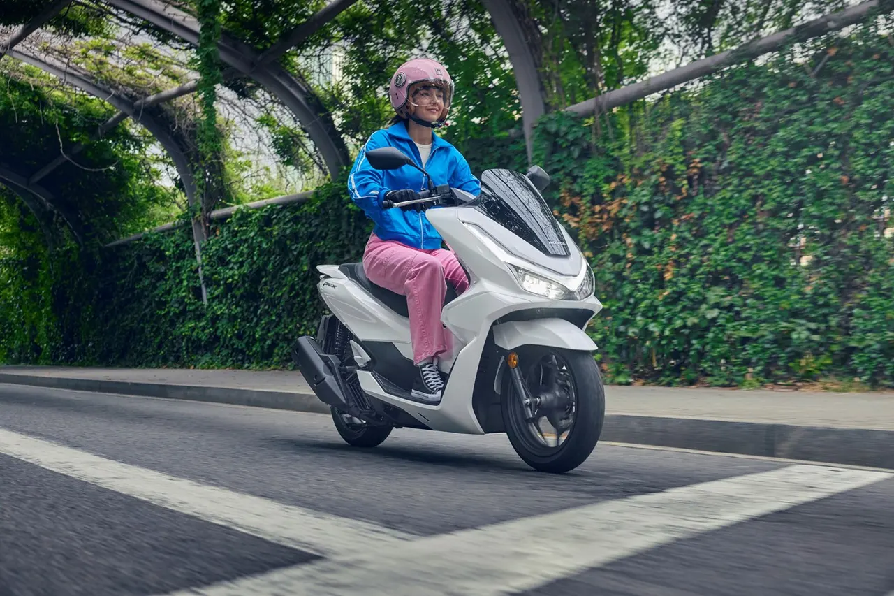 Mujer con casco rosa conduciendo una scooter Honda PCX 125 blanca en una calle arbolada
