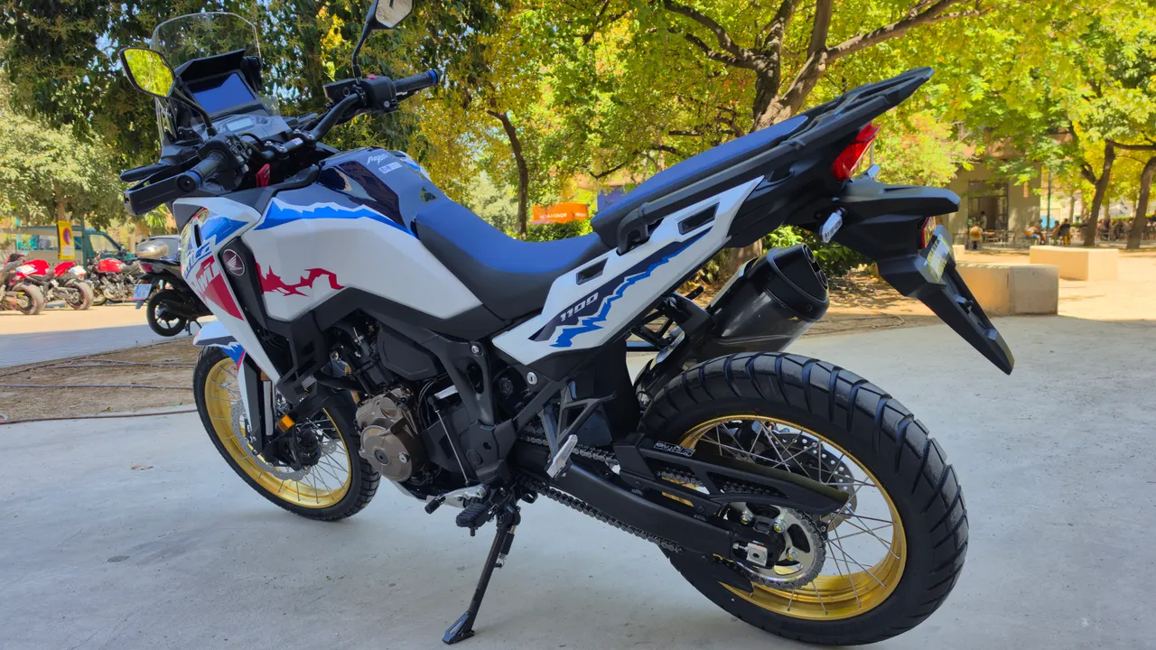 Honda CRF1100L Africa Twin blanca con detalles azules y rojos, ruedas doradas, vista trasera.
