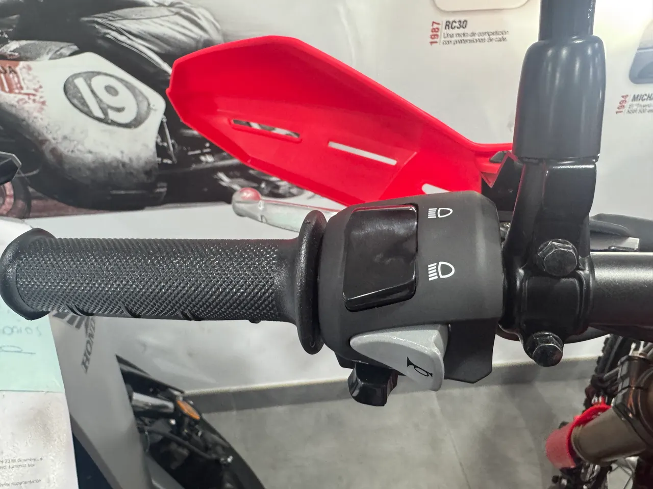 Detalle del manillar izquierdo de una Honda CRF300L con puño, controles de luces y bocina, y protector de mano rojo.