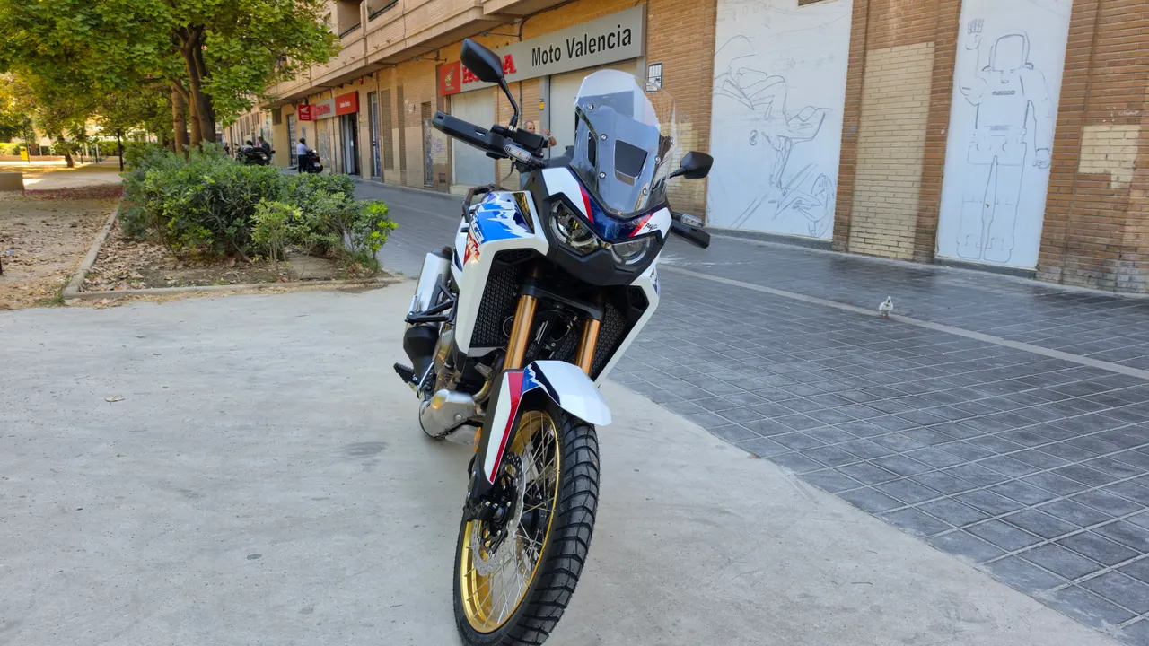 Moto Honda CRF1100L Africa Twin blanca con detalles azules y rojos, y llantas doradas, estacionada en una calle urbana.