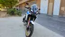 Moto Honda CRF1100L Africa Twin blanca con detalles azules y rojos, y llantas doradas, estacionada en una calle urbana.