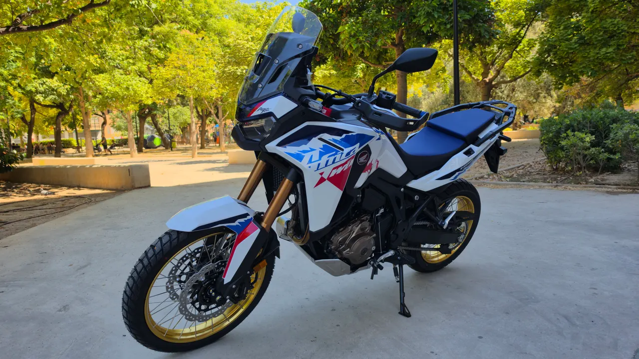 Moto Honda CRF1100L Africa Twin blanca con detalles azules y rojos, ruedas doradas, estacionada en un parque.