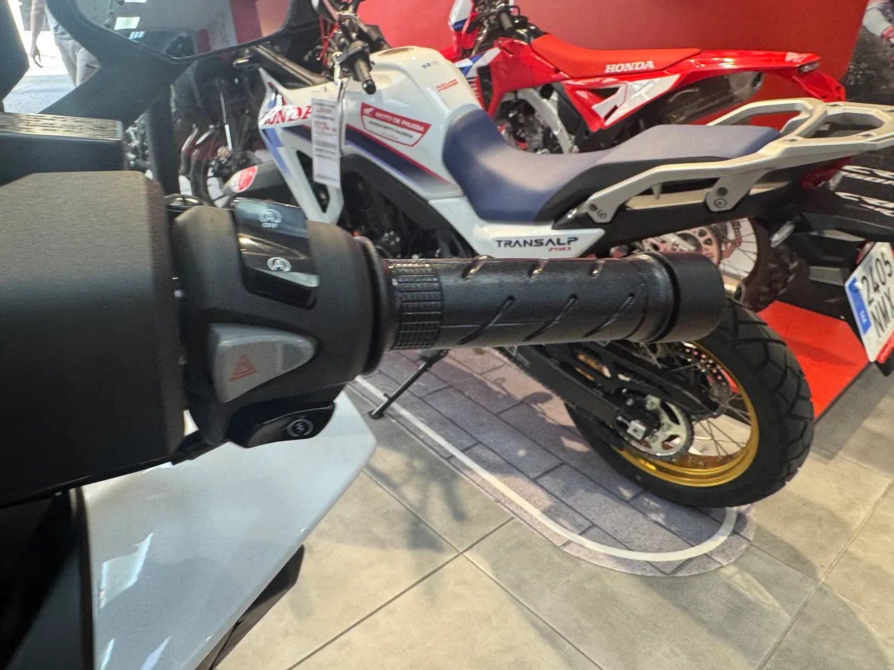 Primer plano del manillar de una Honda Forza 125 blanca con una Honda Transalp blanca, azul y roja en segundo plano.