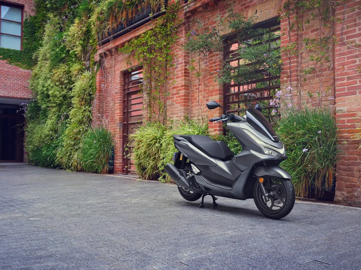 Scooter Honda PCX 125 gris oscuro aparcado frente a una pared de ladrillo con plantas
