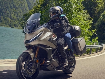 Honda NT1100 2025 Warm Ash Mate Metalizado con dos personas en una carretera frente a un lago, vemos la pantalla en su posicion alta para un mayor confort